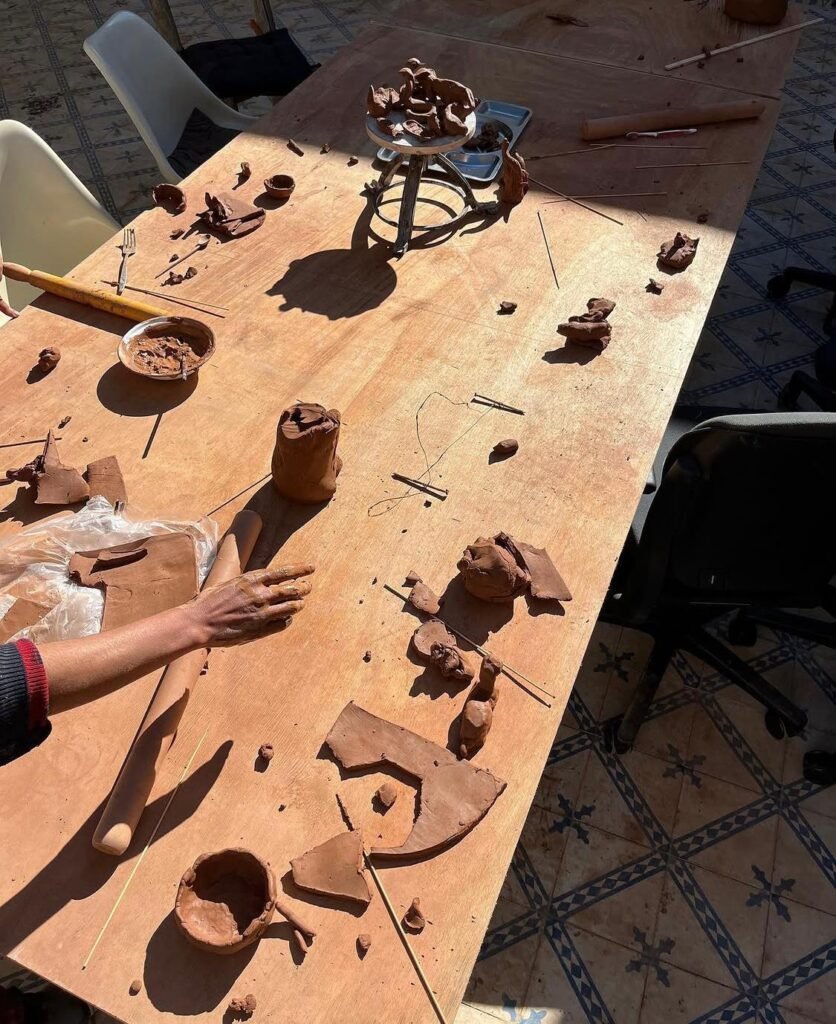 Overhead view of a sunlit table covered with clay scraps, pottery tools, bowls, and a person's hand.