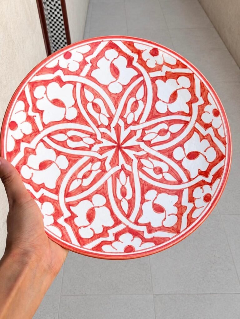 Hand holding a round white decorative plate with red floral and geometric patterns.