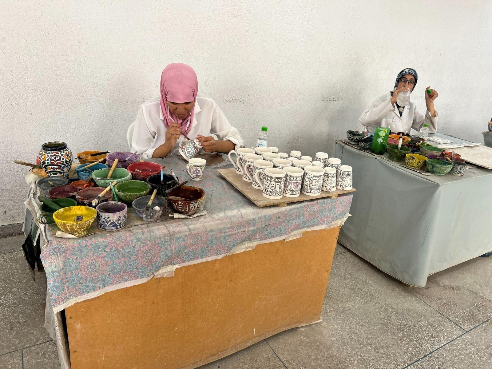 Two women prepare food behind tables with bowls, ingredients, and rows of patterned cups.
